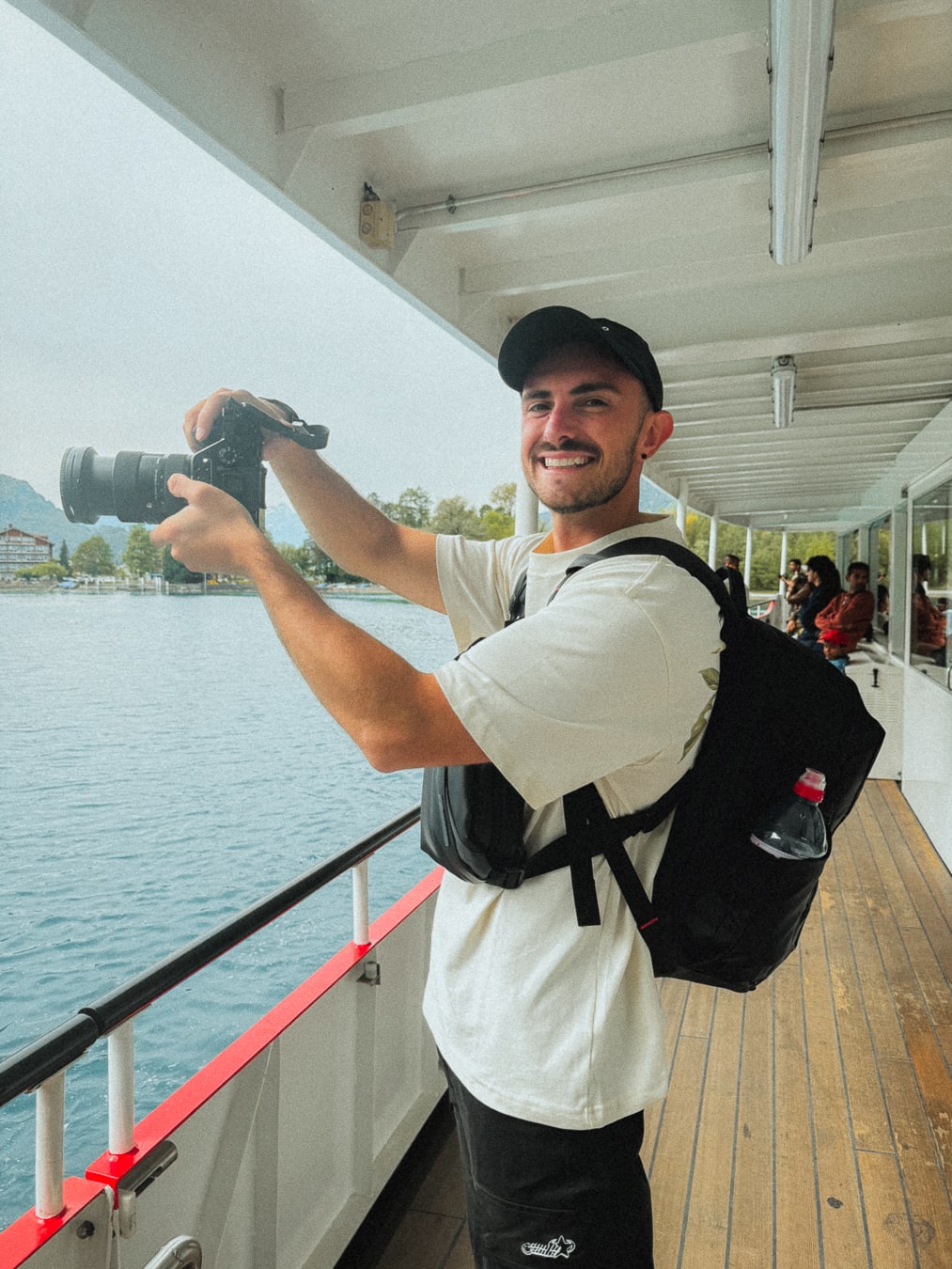 Lachie holding his camera on a boat, with a lake and mountains behind him.